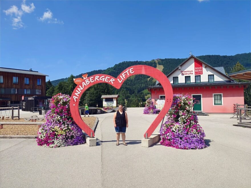 Eine Frau steht in einem großen roten Herz-Schild mit der Aufschrift „ANNABERGER LIFTE“, umgeben von Blumen, im Freien vor Gebäuden und Bergen