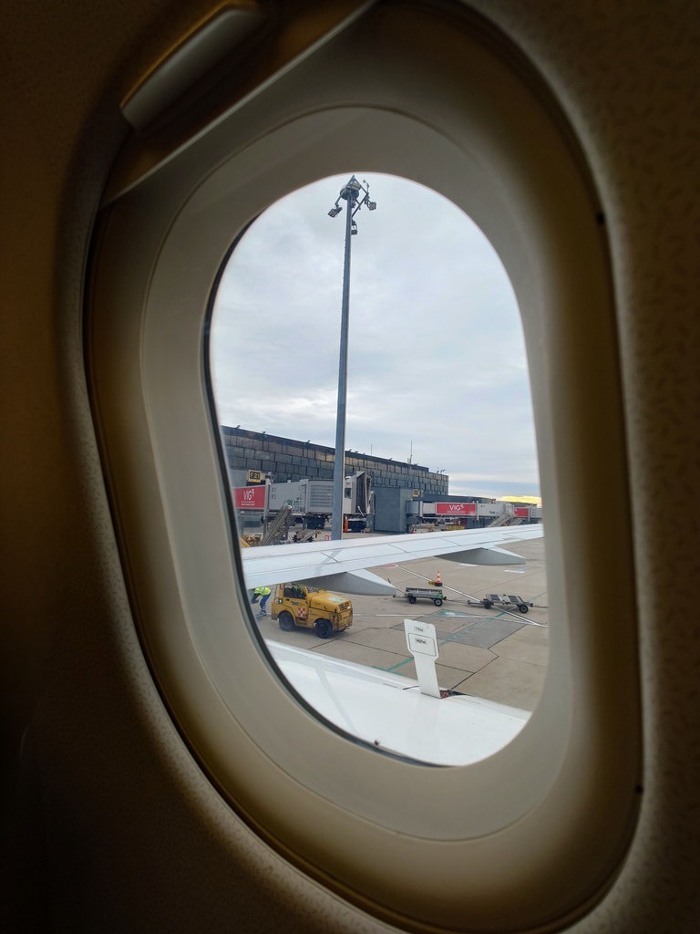 Durch das ovale Fenster ist der Flugzeugflügel zu sehen, ein gelbes Fahrzeug auf dem Vorfeld und das Terminalgebäude im Hintergrund