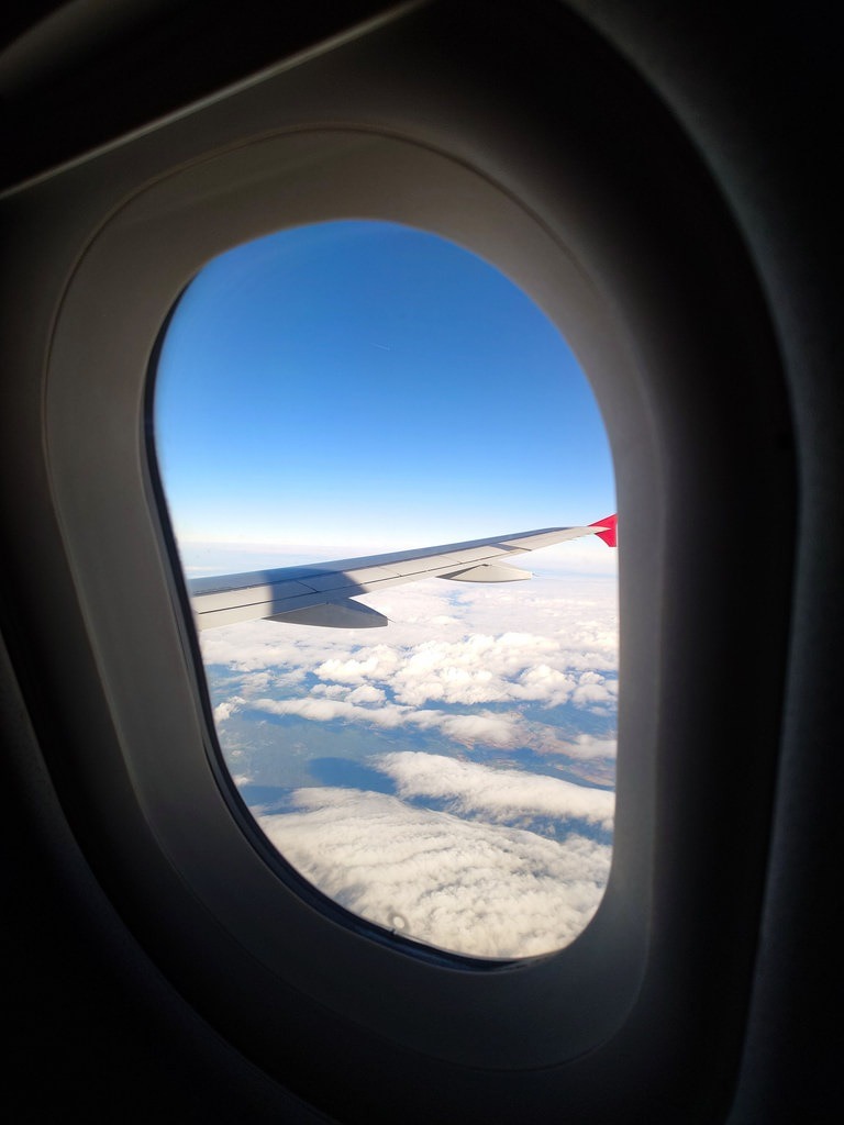 Ansicht aus dem Flugzeugfenster: Der weiße Flügel mit rotem Ende erstreckt sich über einer dichten Schicht weißer Wolken vor einem klaren blauen Himmel