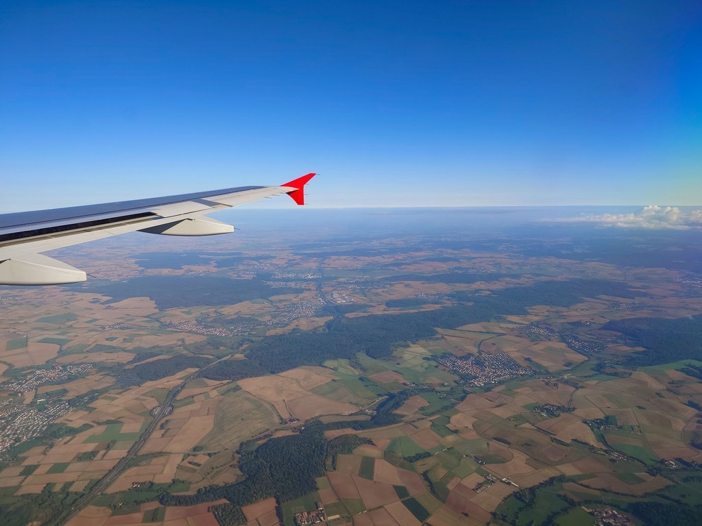 Ein weißer Flugzeugflügel mit roter Flosse ragt in den blauen Himmel über einer Landschaft aus Wäldern und einem Mosaik aus braunen sowie grünen Ackerflächen