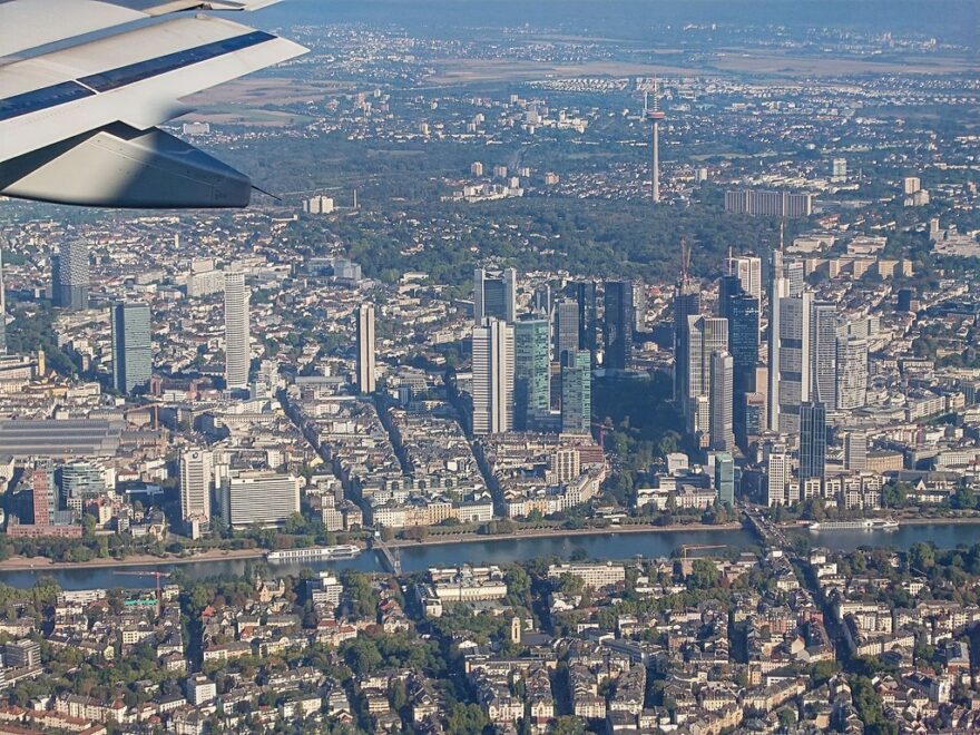 Luftaufnahme einer Stadt mit Wolkenkratzern und Wohngebieten am Fluss, aufgenommen aus einem Flugzeug mit sichtbarem Tragflächenrand oben links