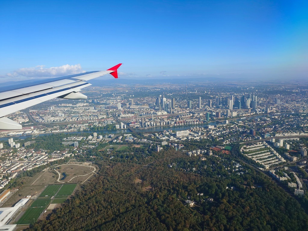 Ein weißer Flugzeugflügel mit rotem Ende ragt links ins Bild. Dahinter liegt eine Stadt mit Hochhäusern, einem Fluss und Wäldern unter klarem Himmel