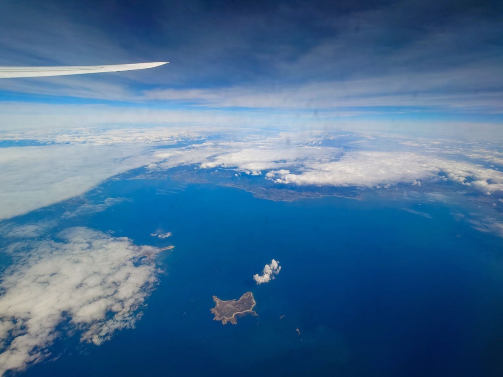 Luftaufnahme, die links oben einen Flugzeugflügel zeigt und darunter das tiefblaue Meer mit kleinen Inseln sowie verstreute weiße Wolken