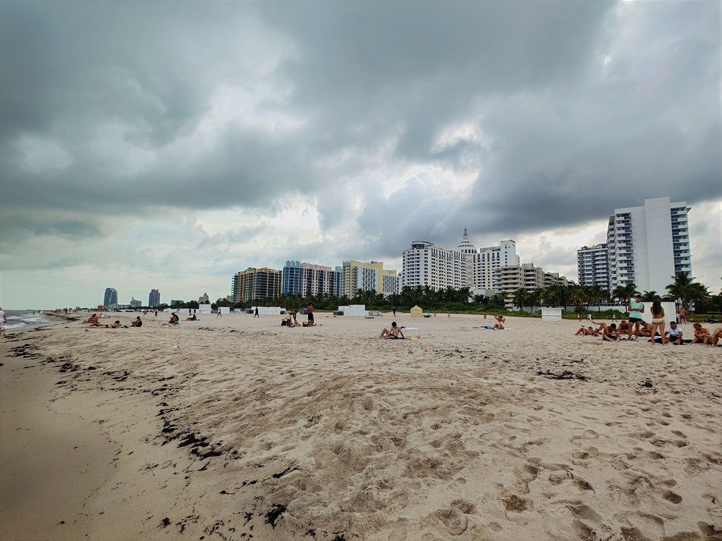 Ein breiter Sandstrand mit zahlreichen Menschen unter einem bewölkten Himmel. Im Hintergrund stehen hohe weiße Gebäude und Palmen, links liegen Algenreste auf dem Sand