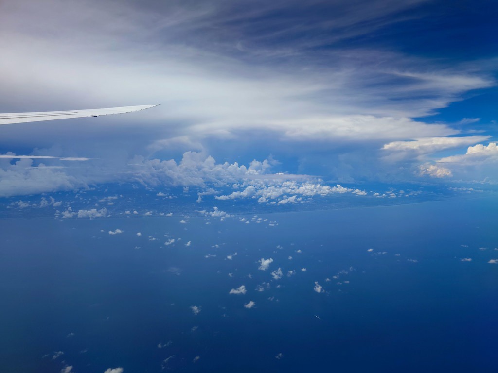 Ein Teil des weißen Flugzeugflügels ist links oben zu sehen, der über einer weiten blauen Wasserfläche und unter einer Schicht weißer Wolken hinwegreicht