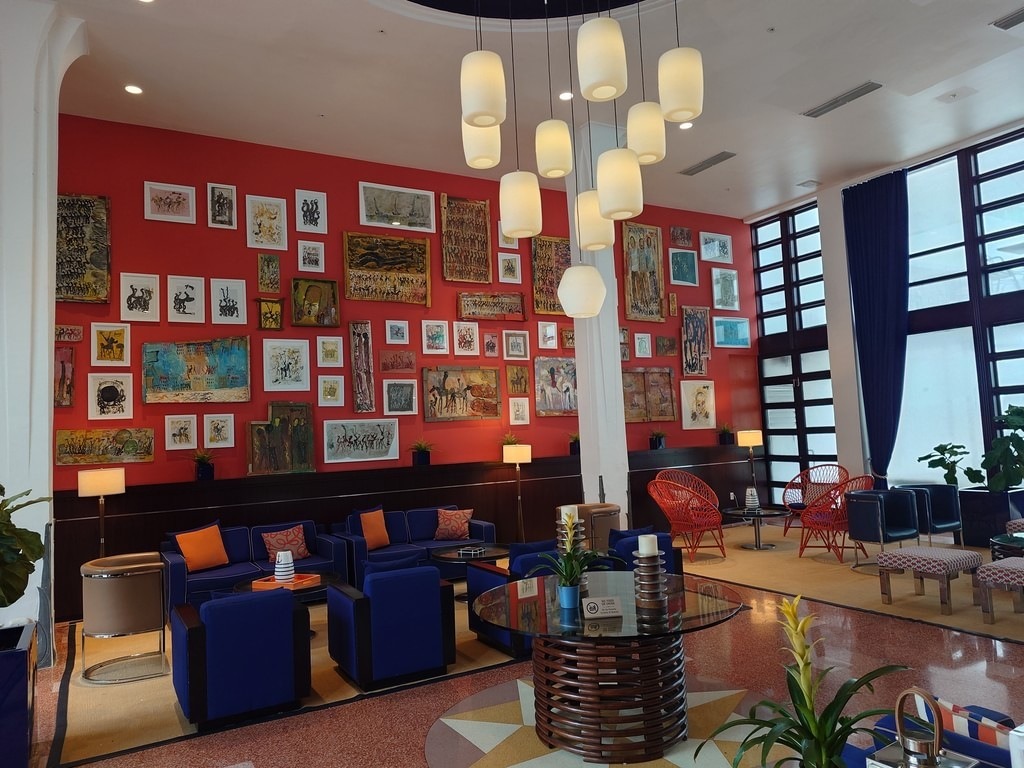 Eine rote Wand voller Bilder und Kunstwerke. Davor stehen blaue Sofas mit orangefarbenen Kissen, Tische und moderne Leuchten. Rechts hohe Fenster mit dunklen Vorhängen