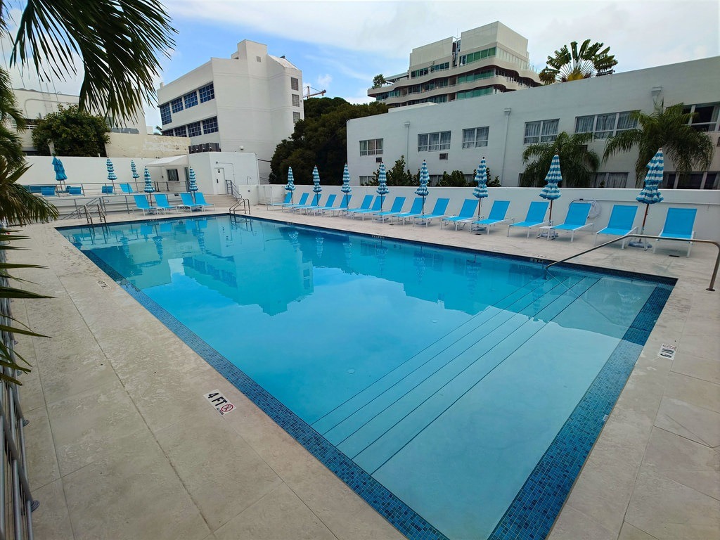 Ein rechteckiger Pool mit blauem Wasser und Kachelrand, umgeben von leeren blauen Sonnenliegen und geschlossenen, gestreiften Sonnenschirmen vor weißen Gebäuden und Palmen
