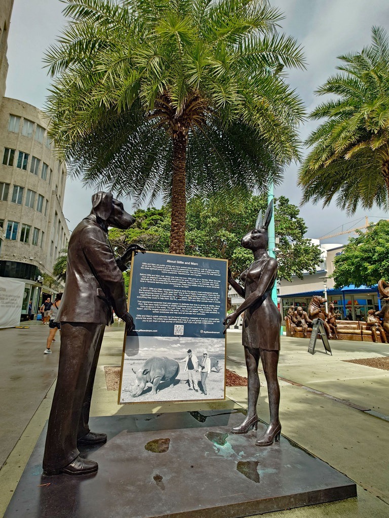 Eine bronzene Hundestatue in Anzug und eine Kaninchenfigur in Kleid halten ein Text- und Bildschild. Im Hintergrund sind Palmen und ein helles Gebäude zu sehen