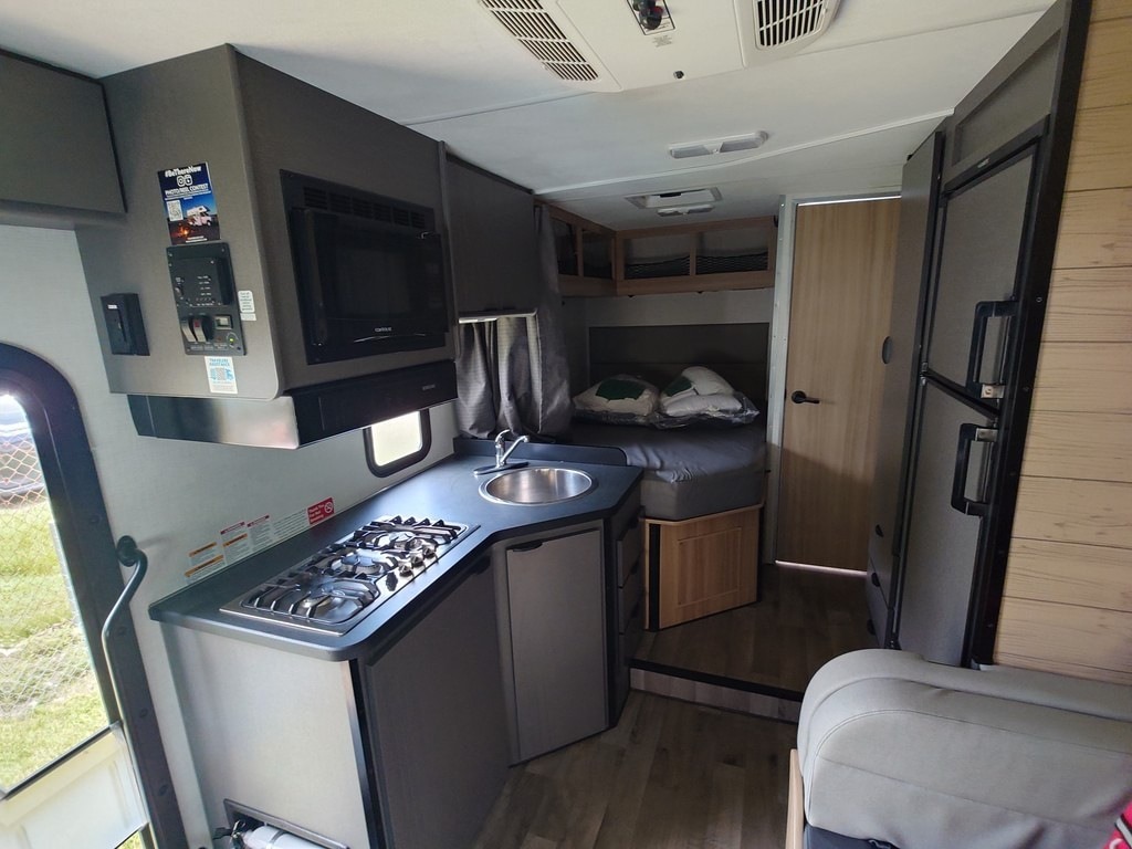 Der Innenraum eines Wohnmobils zeigt links die Küche mit Herd, Spüle und Mikrowelle. Weiter hinten befindet sich ein Bett, rechts ein Kühlschrank