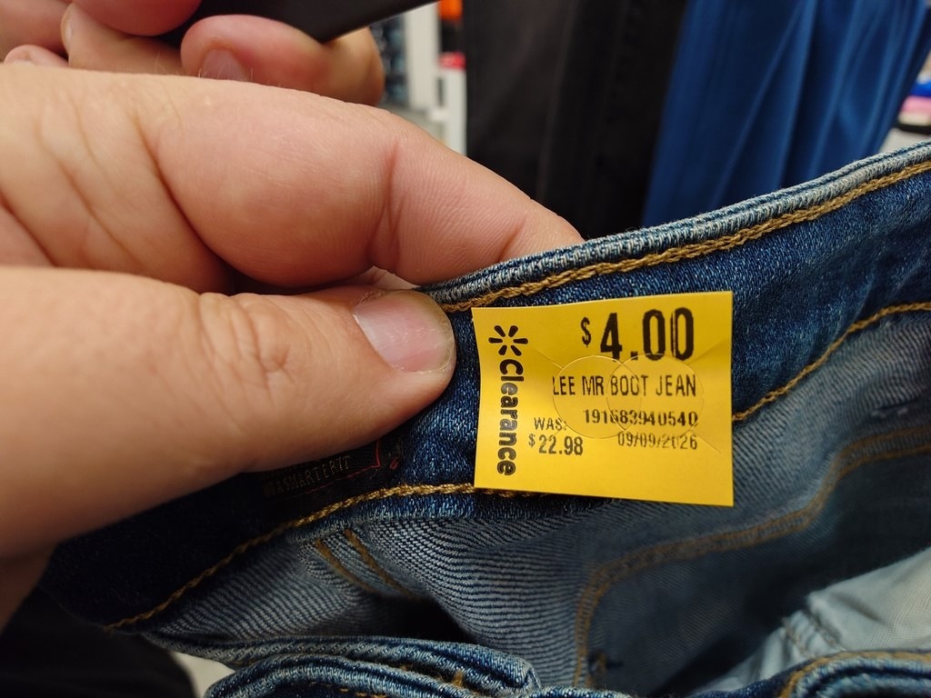 Eine Hand hält den Bund einer Jeans hoch, um ein gelbes Preisetikett zu zeigen. Es listet "Lee MR Boot Jean" für 4,00 Dollar, vormals 22,98 Dollar