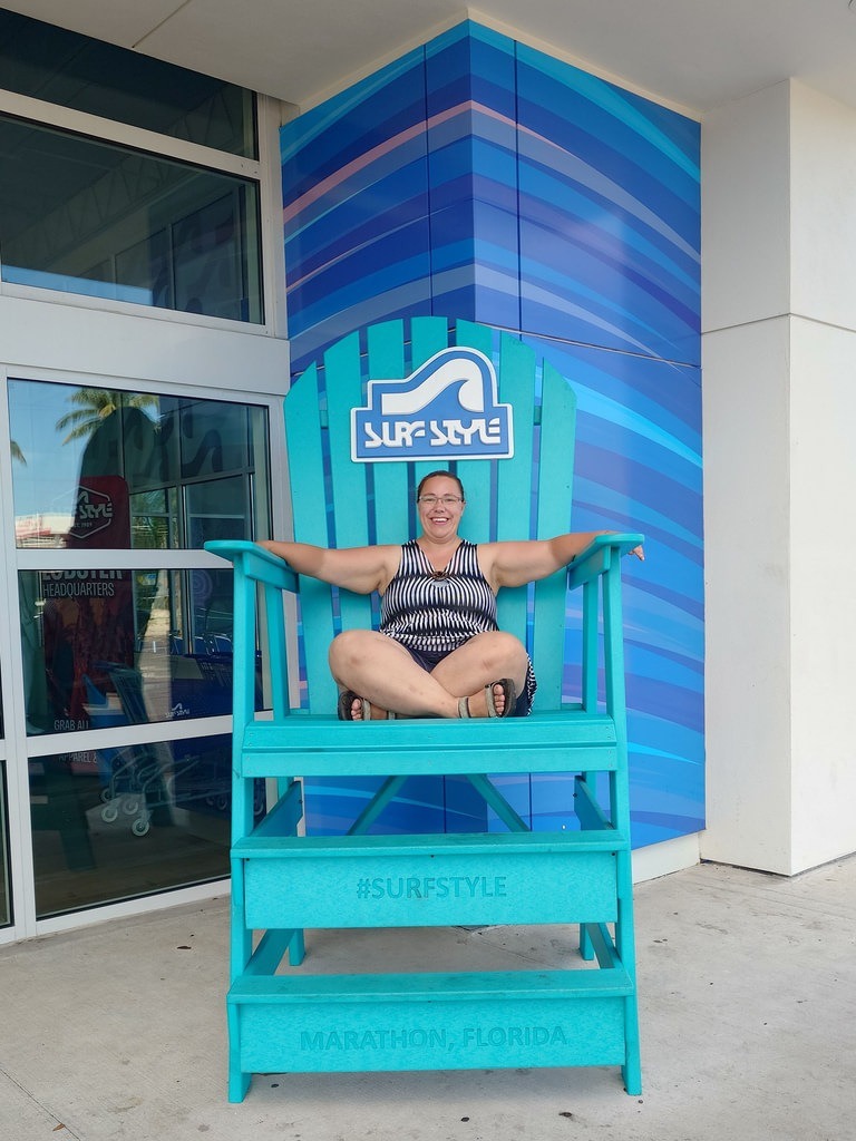 Eine lächelnde Frau sitzt auf einem übergroßen türkisfarbenen Liegestuhl mit den Aufschriften „Surf-Style“ und „Marathon, Florida“