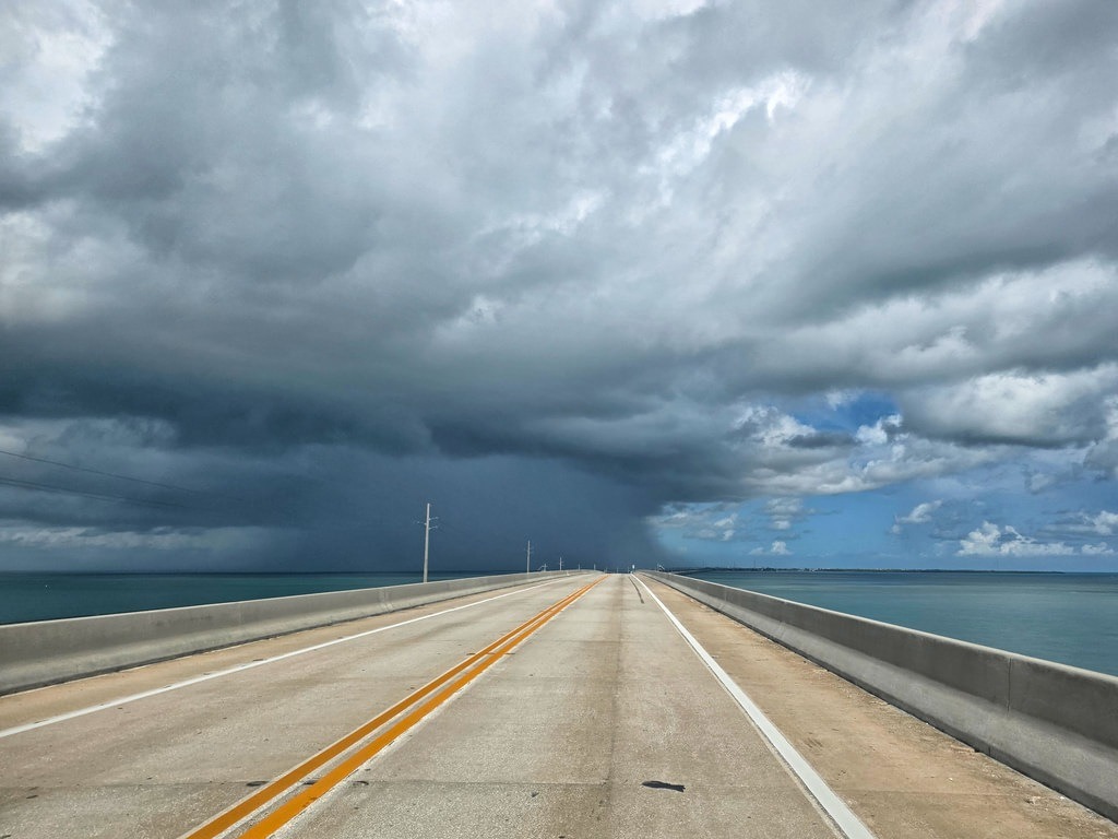 Zweispurige Straße auf einer Brücke führt über das Wasser, während am Horizont dunkle Gewitterwolken aufziehen und die Landschaft bedecken