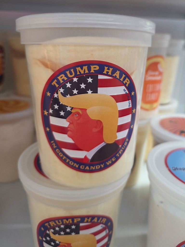 Ein Behälter mit hellgelber Zuckerwatte zeigt einen Aufkleber mit Donald Trumps Profil, US-Flaggen und den Texten „Trump Hair“ sowie „In Cotton Candy We Trust“