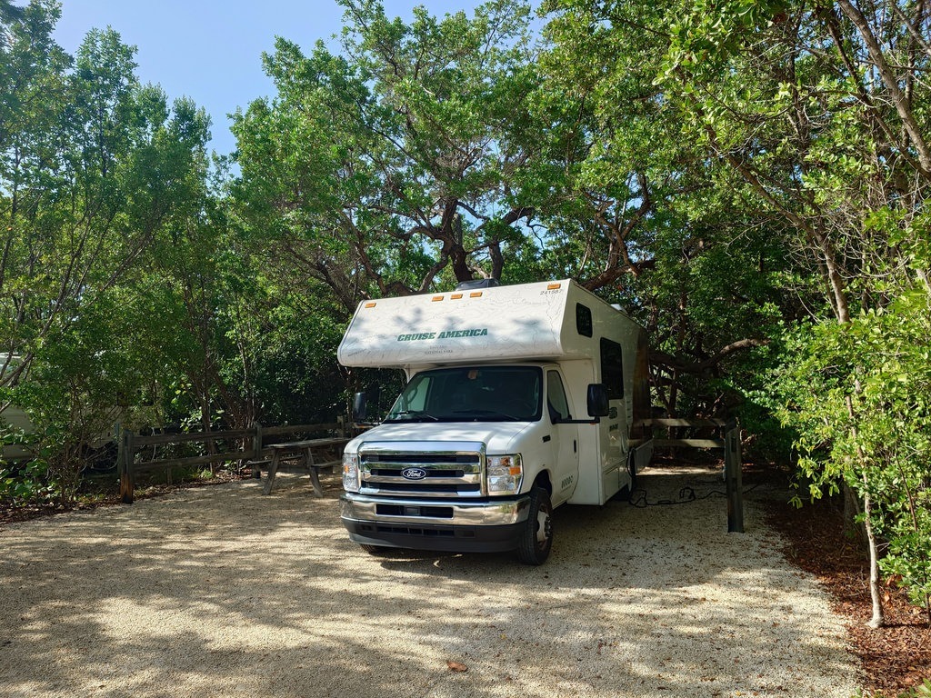 Ein weißes Cruise America Wohnmobil steht auf einer kiesigen Fläche, umgeben von Bäumen und Sträuchern an einem sonnigen Tag