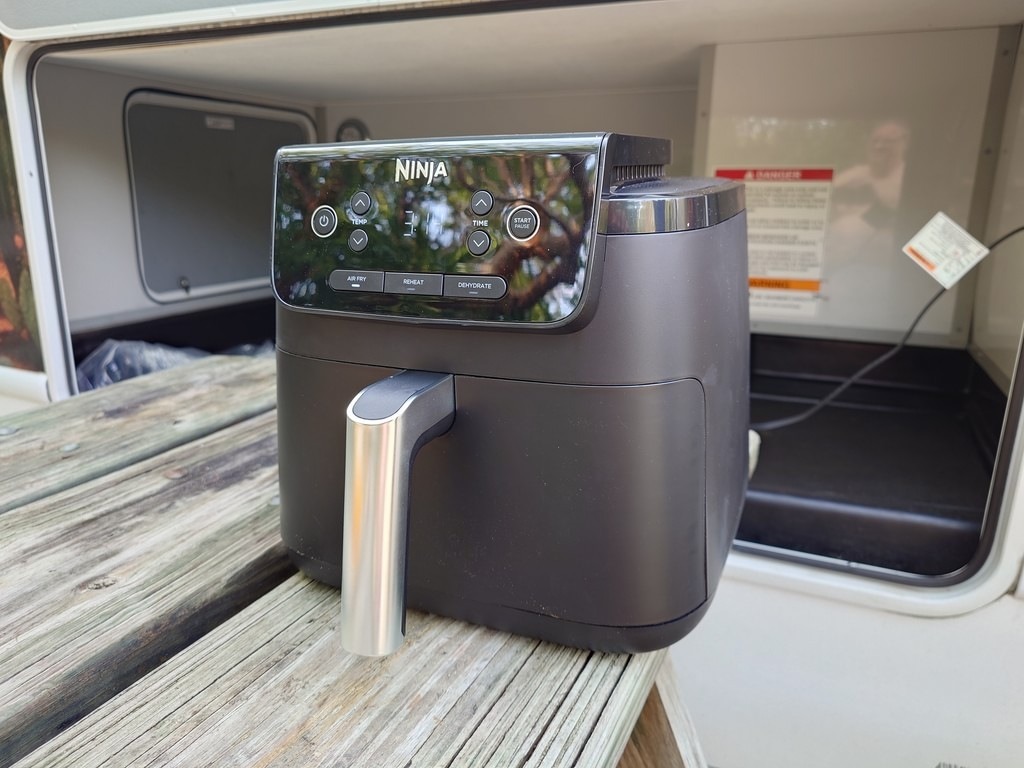 Dunkelgrauer Ninja Air Fryer mit Display und silbernem Griff auf einer Holzplatte. Im Hintergrund sind weiße Wände und ein Warnschild zu erkennen