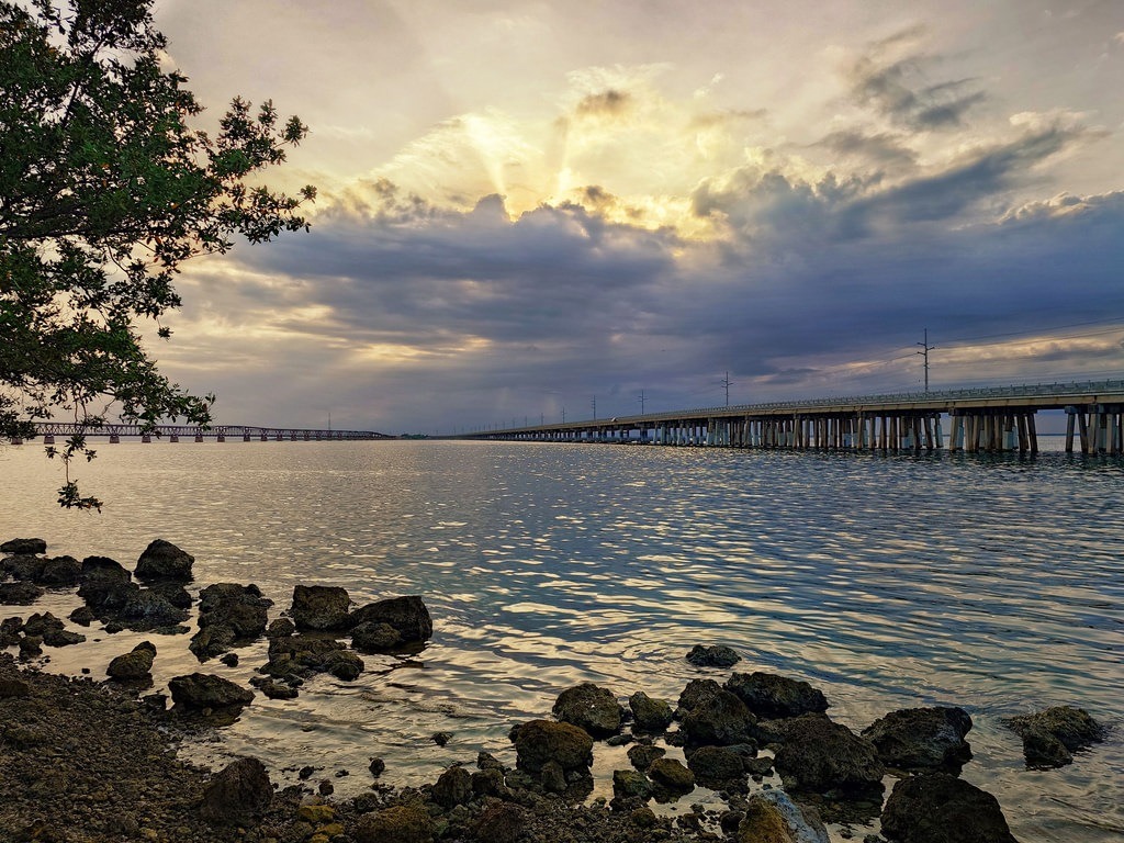 Unter einem bewölkten Himmel brechen Lichtstrahlen hindurch, während eine lange Brücke über das Wasser führt und im Vordergrund Felsen sowie ein Baum zu sehen sind