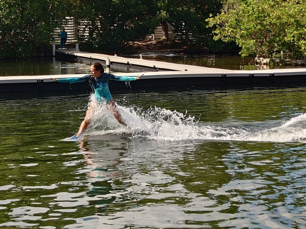 Eine Person in einem blaugrünen Oberteil balanciert auf einem Wakeboard und wirbelt weißes Gischt auf, im Hintergrund ist ein langer Holzsteg vor Bäumen zu sehen