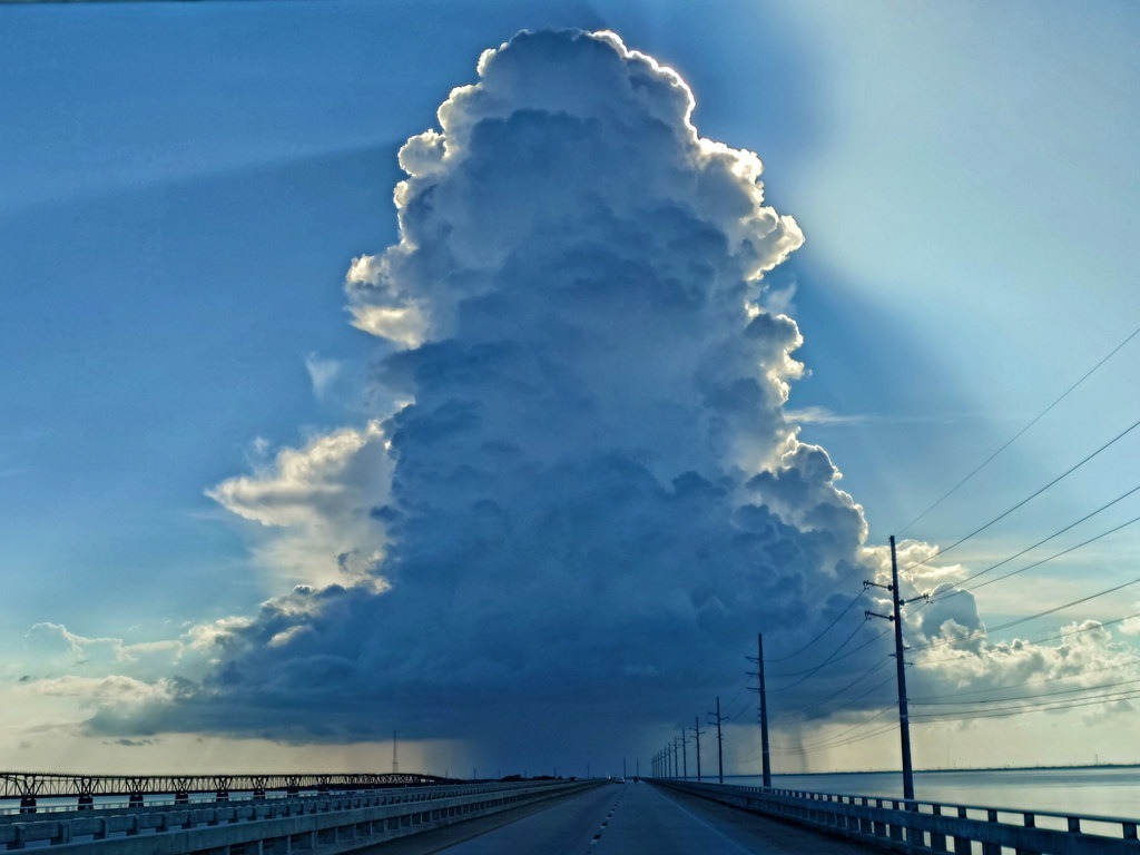 Eine asphaltierte Brücke führt in die Ferne, dominiert von einer riesigen, türmenden Wolke. Rechts verlaufen Strommasten parallel zur Fahrbahn