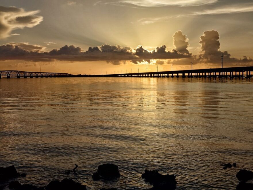 Eine lange Brücke erstreckt sich dunkel über das Wasser. Felsen ragen im Vordergrund aus dem spiegelnden Gewässer, während die Sonne hinter Wolken scheint