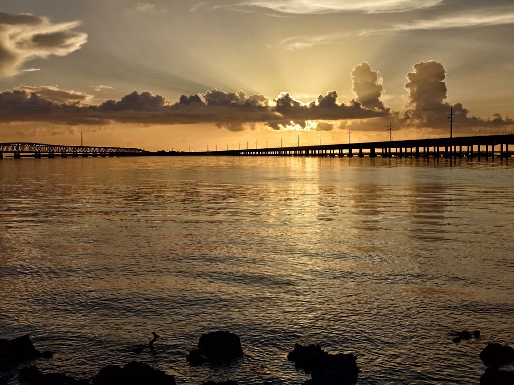Eine lange Brücke erstreckt sich dunkel über das Wasser. Felsen ragen im Vordergrund aus dem spiegelnden Gewässer, während die Sonne hinter Wolken scheint