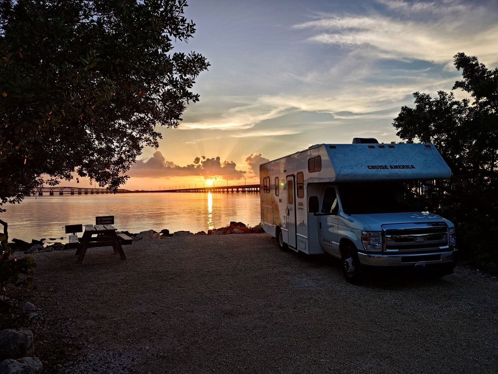 Ein Cruise America Wohnmobil steht am Ufer. Im Hintergrund leuchtet die Sonne über einer Brücke. Links steht ein Picknicktisch unter einem Baum