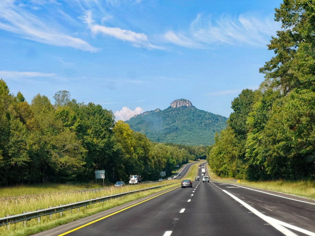 Ein Asphaltweg führt durch grüne Bäume hindurch direkt auf einen Berg mit felsigem Gipfel zu, während einige Autos die Straße befahren