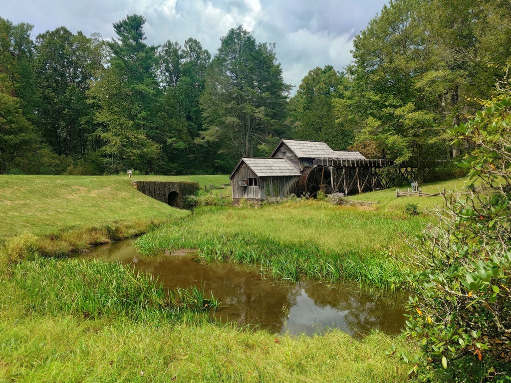 Eine hölzerne Mühle mit großem Wasserrad steht am Ufer eines Baches, umgeben von grüner Wiese und einem dichten Wald unter bewölktem Himmel