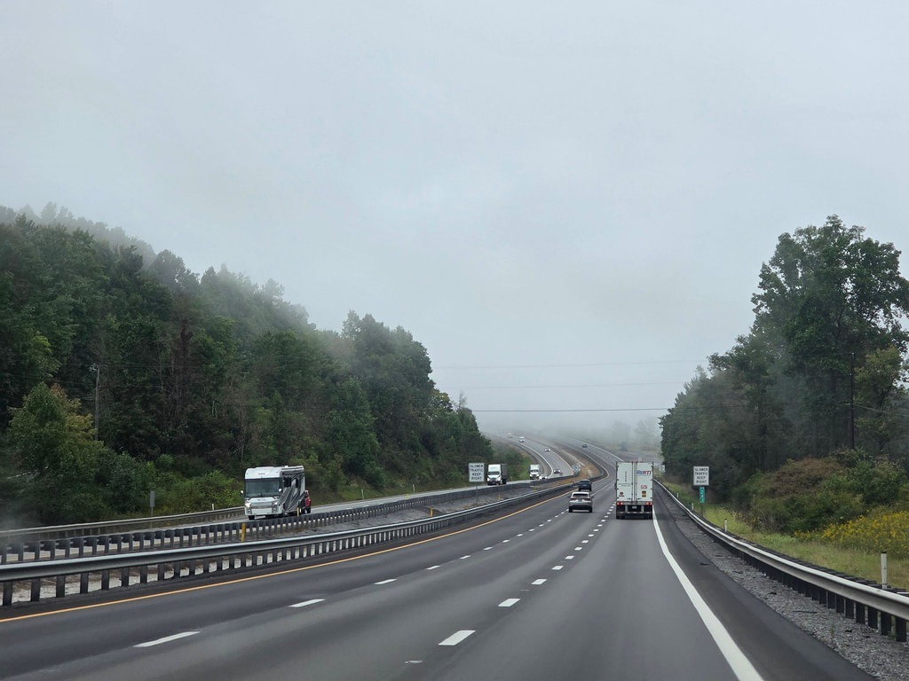 Unter dem nebligen Himmel verläuft eine feuchte Autobahn durch Waldgebiete. Links steht ein weißes Wohnmobil, rechts fahren Lastwagen und Autos
