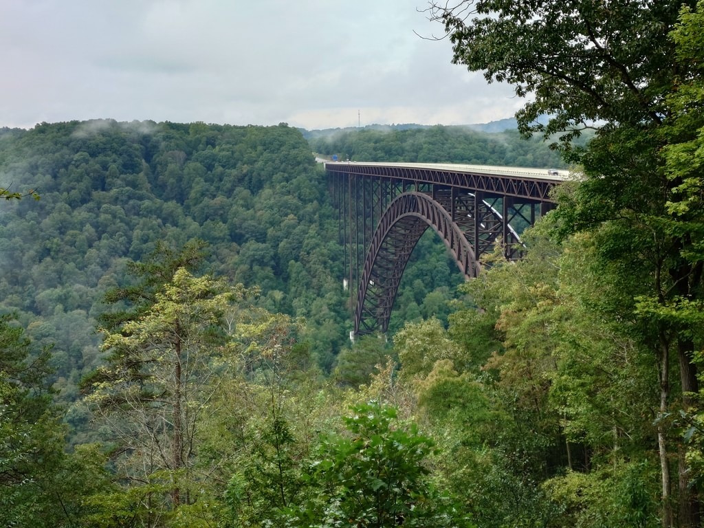 Eine massive Stahlbogenbrücke überspannt ein tiefes, dicht bewaldetes Tal unter einem bewölkten Himmel. Im Hintergrund sind neblige Hügel zu erkennen