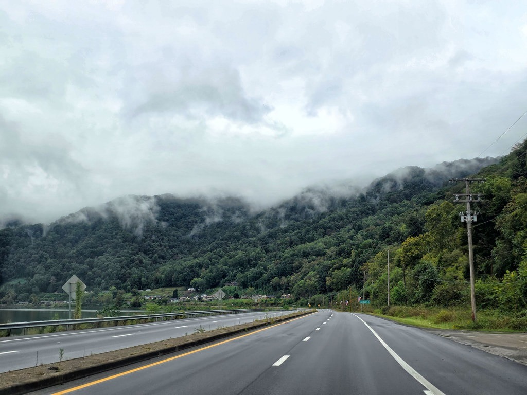 Eine asphaltierte Straße verläuft parallel zu einem Gewässer in Richtung auf bewaldete Berge, deren Gipfel in tief hängenden Wolken verschwinden