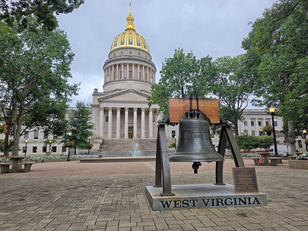 Eine Bronzeglocke auf einem Gestell trägt die Aufschrift „WEST VIRGINIA“. Im Hintergrund steht ein weißes Gebäude mit einer goldenen Kuppel