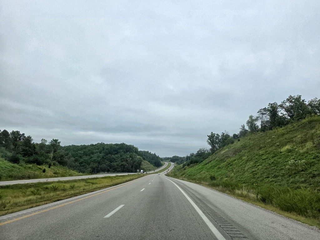 Eine mehrspurige Autobahn windet sich unter bewölktem Himmel sanft nach rechts. Grüne Wiesen und Bäume säumen den Weg, auf dem wenige Fahrzeuge zu sehen sind