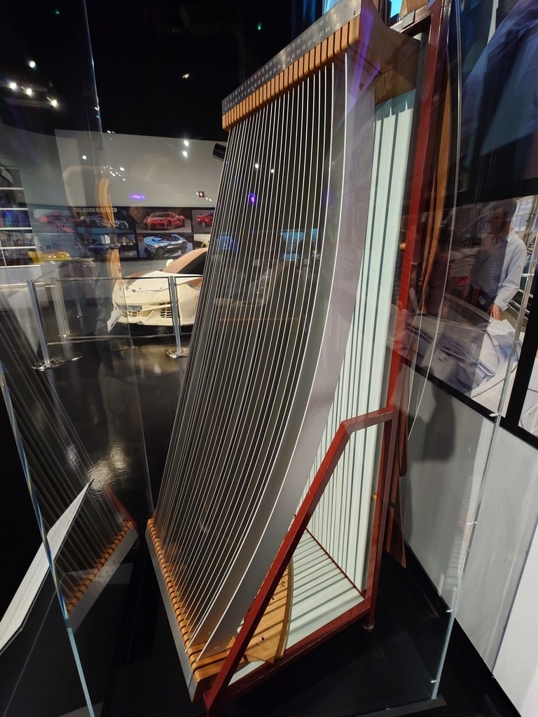 Ein massiver Rechenschieber mit vertikalen Metallstäben steht in einem hellen Museumsraum vor einer Wand mit Autoelementen und Skizzen, während sich die Umgebung in einer gläsernen Vitrine spiegelt