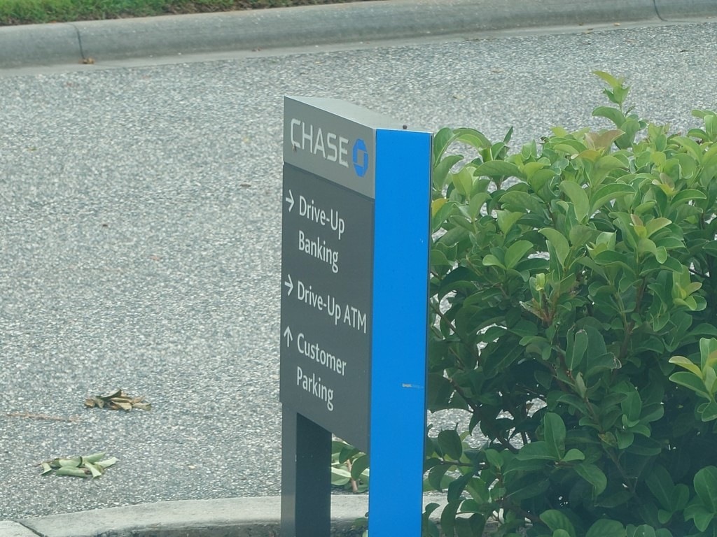Graufarbenes Chase-Bank-Wegweiser-Schild mit blauer Akzentleiste und weißer Beschriftung für Drive-Up Banking, Drive-Up ATM und Customer Parking, aufgestellt an einem asphaltierten Parkplatz neben sattgrünen Sträuchern