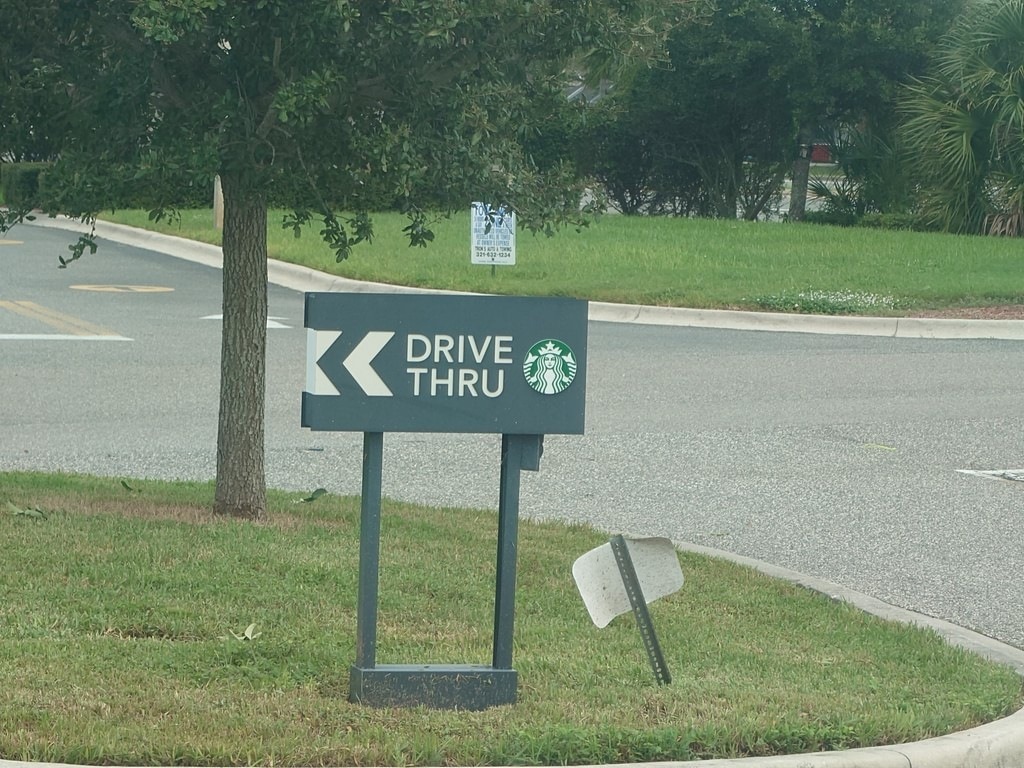 Dunkelgraues Hinweisschild mit weisser Aufschrift „DRIVE THRU“ und dem runden grünen Starbucks-Logo, aufgestellt auf einer Verkehrsinsel mit grünem Rasen neben einer asphaltierten Strasse und Bäumen im Hintergrund