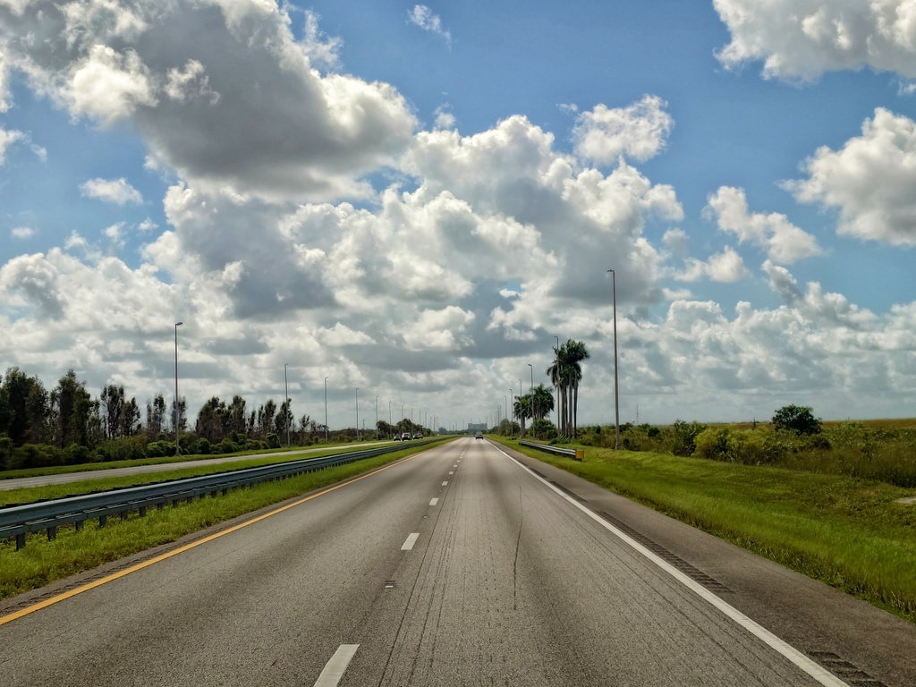 Eine breite Asphalt-Autobahn führt durch die flache Sumpflandschaft Floridas, gesäumt von grasbewachsenen Rändern, einzelnen Palmen und weit entfernten Baumgruppen unter einem hellblauen Himmel mit dichten, weissen Cumuluswolken