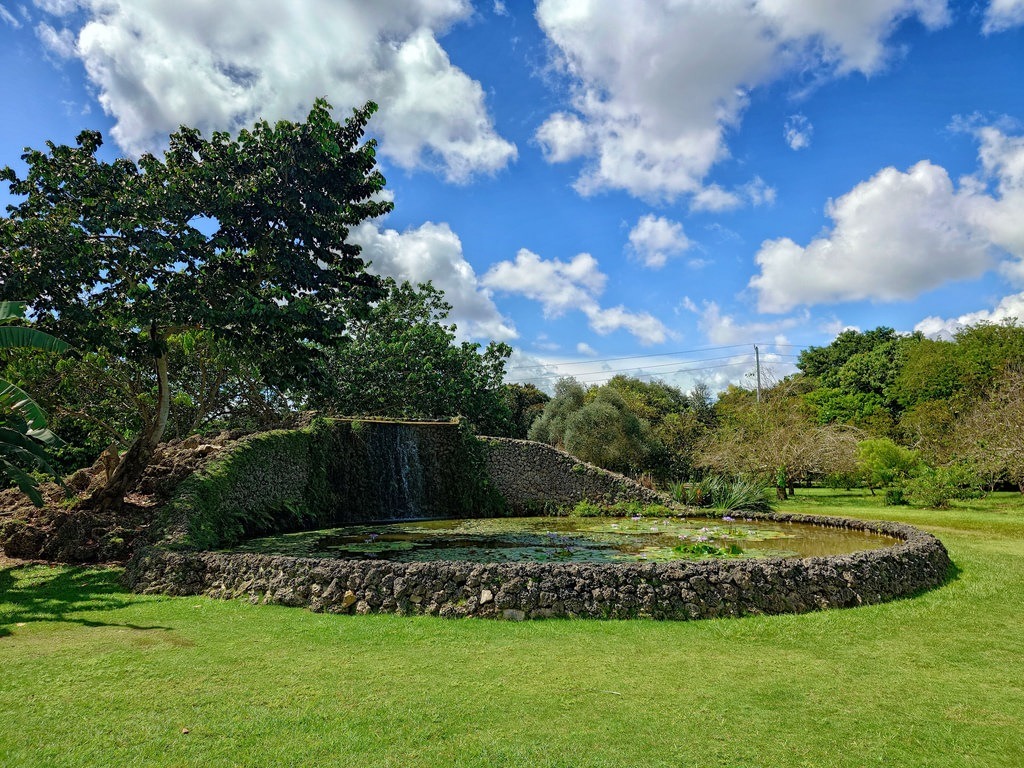 Ein runder Natursteinteich mit Seerosen und einem kleinen künstlichen Wasserfall, eingebettet in eine sattgrüne Wiesenlandschaft unter einem leuchtend blauen Himmel mit weissen Wolken