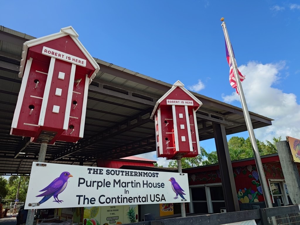 Zwei rot-weisse Mehrfamilien-Nistkästen mit der Aufschrift „ROBERT IS HERE“ und ein Hinweisschild „The Southernmost Purple Martin House in The Continental USA“ unter einem dunklen Vordach vor blauem Himmel mit weissen Wolken und einer US-Flagge am Mast