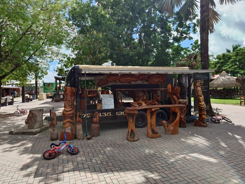 Ein umgebauter schwarzer Verkaufshänger mit kunstvollen Holzskulpturen und Tiki-Figuren steht auf einem gepflasterten Platz im Schatten hoher Laubbäume und Palmen, davor parken zwei kleine Fahrräder