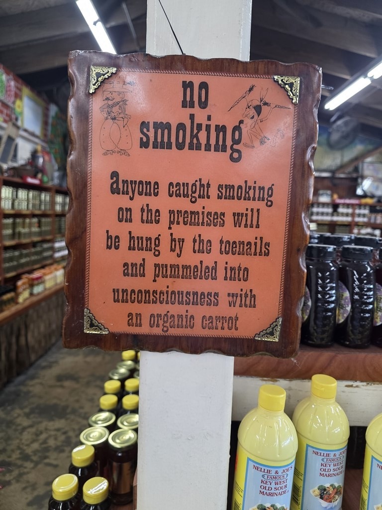 Ein orangefarbenes Hinweisschild mit braunem Holzrahmen und dekorativen Eckbeschlägen hängt an einem weißen Holzpfosten in einem Geschäft, flankiert von Glasgläsern und gelben Flaschen. Der Text lautet: „no smoking. Anyone caught smoking on the premises will be hung by the toenails and pummeled into unconsciousness with an organic carrot“, verziert mit kleinen Comic-Zeichnungen in den Ecken