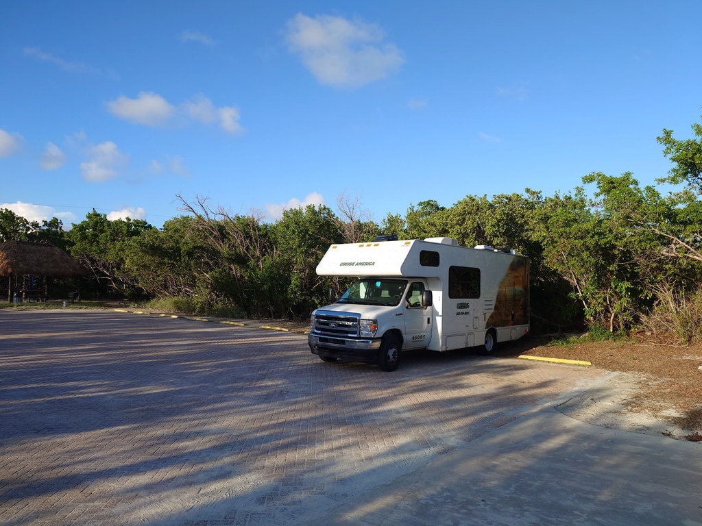 Weisses Wohnmobil von Cruise America mit Aufdruck parkt auf einem hell gepflasterten Parkplatz, im Hintergrund dichte grüne Bäume und ein blauer Himmel mit vereinzelten Wolken