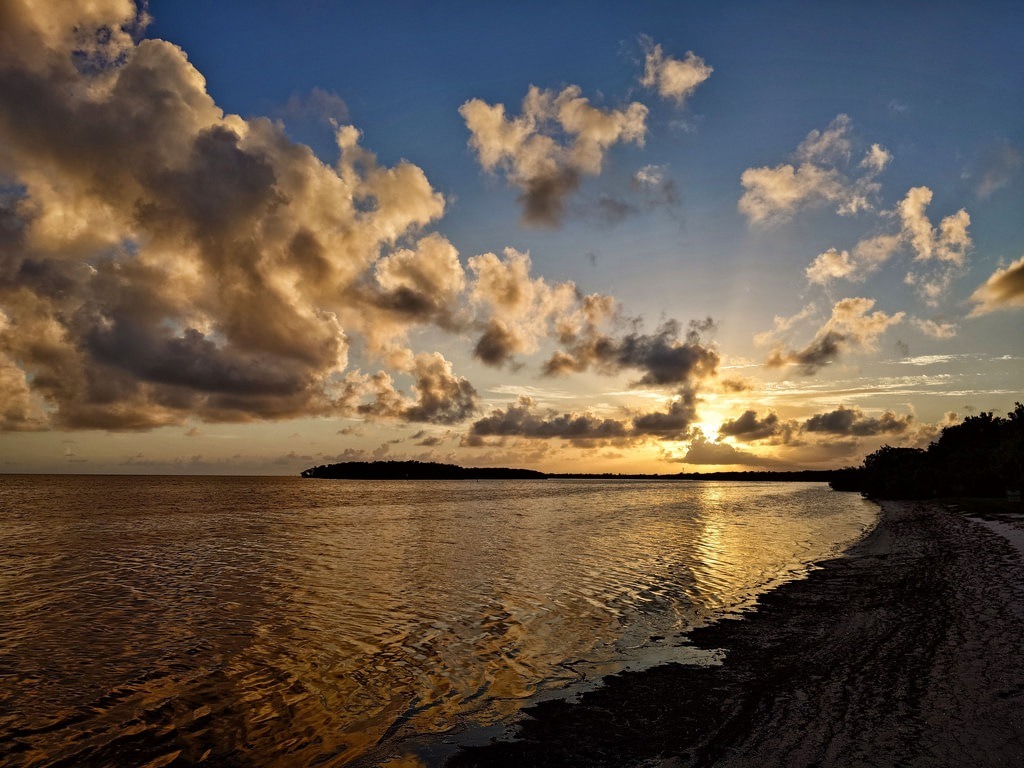 Tropischer Sonnenuntergang an einer Küste mit dunkelgolden schimmerndem Wasser, dramatischen, von der Abendsonne gold- und weißgefärbten Wolkenformationen am tiefblauen Himmel und einer flachen, bewaldeten Landzunge im Hintergrund