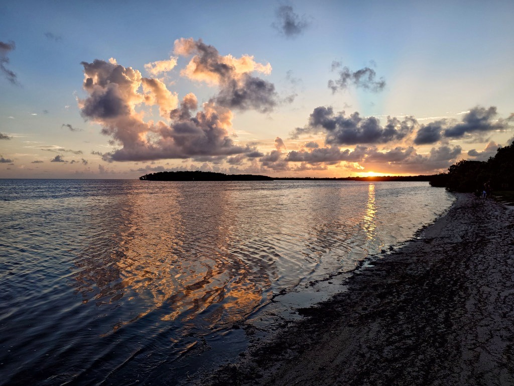 Stimmungsvoller Sonnenuntergang über einer ruhigen Bucht mit spiegelndem Wasser, golden leuchtenden Wolkenformationen, dunklen Waldumrissen am Horizont und einem naturbelassenen Ufer im Vordergrund
