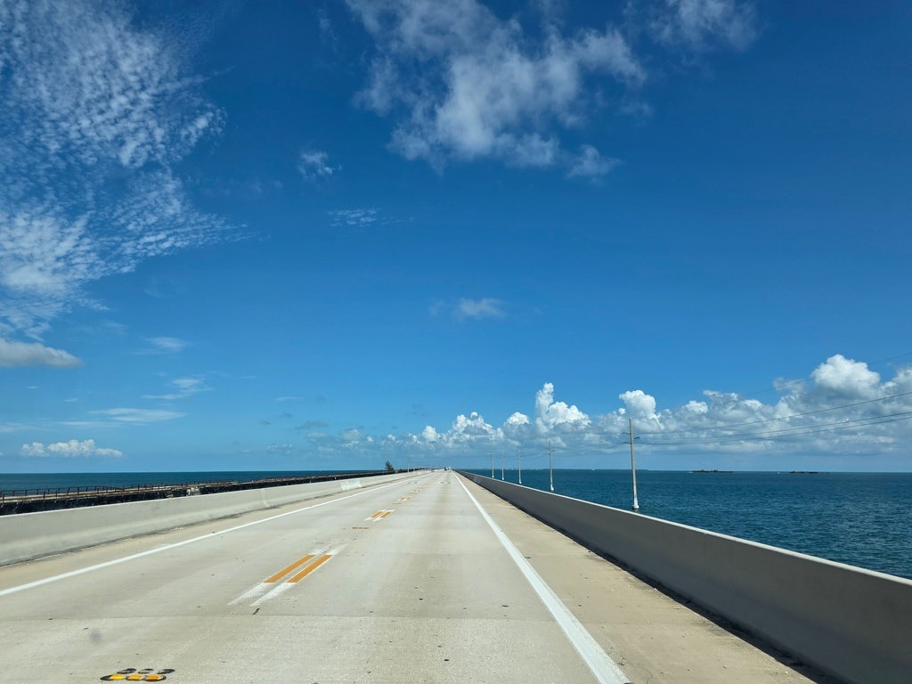 Die hellgraue Fahrbahn der Seven Mile Bridge führt geradeaus durch das tiefblaue Ozeanwasser der Florida Keys, flankiert von weißen Fahrbahnmarkierungen, gelben Mittelstreifen und niedrigen Betonbegrenzungen unter einem weiten, leuchtend blauen Himmel mit feinen weißen Wolkenfetzen am Horizont