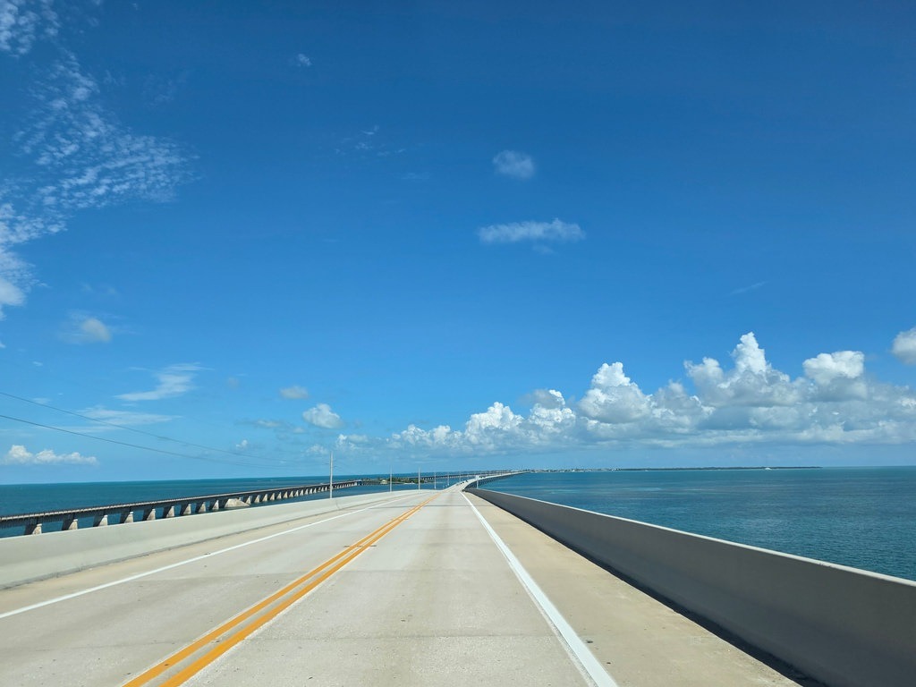 Die hellgraue Fahrbahn der Overseas Highway verläuft schnurgerade mit doppelter gelber Mittelmarkierung über das tiefblaue Ozeanwasser der Florida Keys, begleitet von der historischen Eisenbahnbrücke und einem weiten, hellblauen Himmel voller kleiner Quellwolken