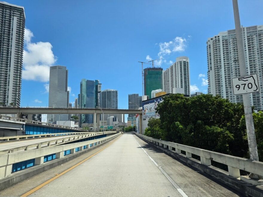 Blick auf eine leicht ansteigende Autobahnbrücke der Florida State Road 970 mit hellgrauem Beton und weisser Fahrbahnmarkierung, eingerahmt von dichten grünen Bäumen und einer imposanten Skyline mit modernen Wolkenkratzern und Baukränen unter strahlend blauem Himmel mit leichten Wolken