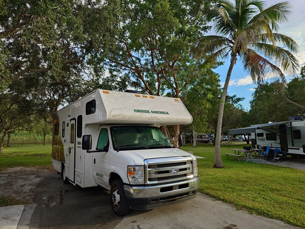 Ein weisses Wohnmobil der Marke Cruise America mit glänzendem Chromkühlergrill steht auf einem asphaltierten Stellplatz im Schatten hoher Laubbäume und einer schlanken Palme, während im Hintergrund weitere Campingfahrzeuge und saftig grüne Wiesen zu sehen sind