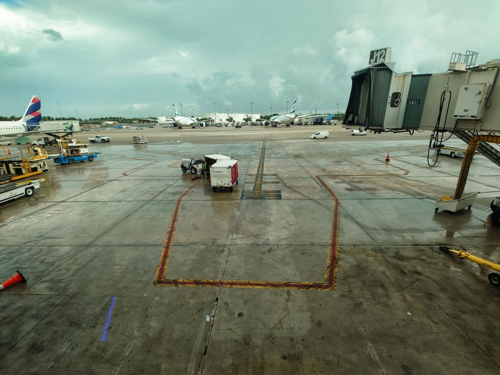 Flugfeld auf einem nassen Rollfeld an Gate J12 mit grauen Betonplatten, mehreren Passagiermaschinen im Hintergrund, einer weissen Fluggastbrücke im Vordergrund und dramatischem Wolkenhimmel