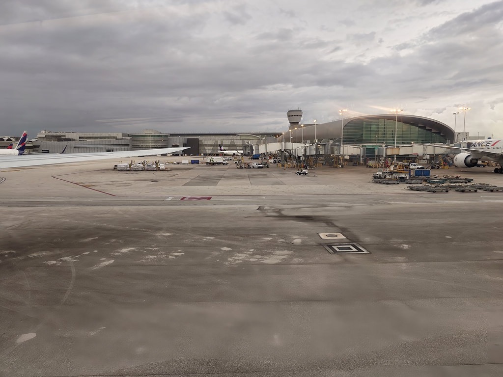 Blick aus dem Flugzeugfenster auf das weite Rollfeld eines grossen Flughafens unter einem dichten, grauen Wolkenhimmel. Im Hintergrund stehen Passagiermaschinen an den beleuchteten Fluggastbrücken des modernen Terminalgebäudes mit grossen Glasfronten und einem markanten Bogendach