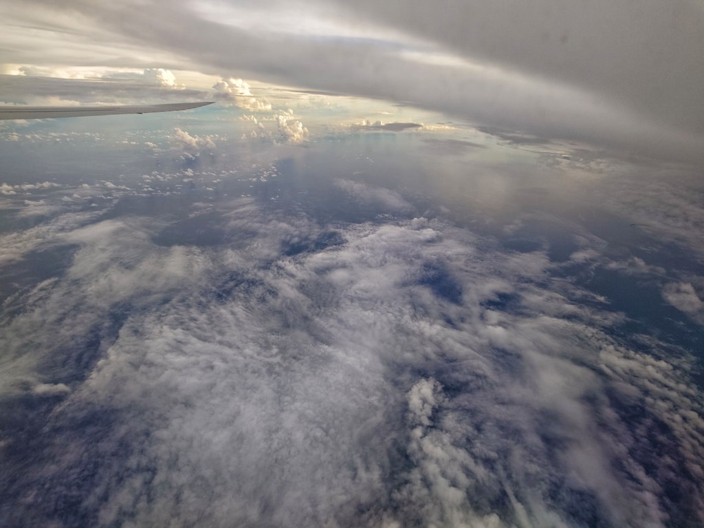 Flügelspitze eines Passagierflugzeugs im linken oberen Bildrand vor einem weiten Ozean aus dichten, weiß-grauen Wolkenfeldern, die sich unter einem dramatischen, teilweisen bedeckten Abendhimmel erstrecken